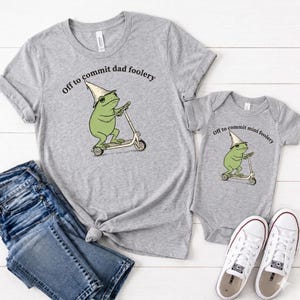 May include: A matching grey t-shirt and baby onesie set featuring a cartoon frog on a scooter. The frog wears a wizard hat. The t-shirt says "Off to commit dad foolery", the onesie says "Off to commit mini foolery."