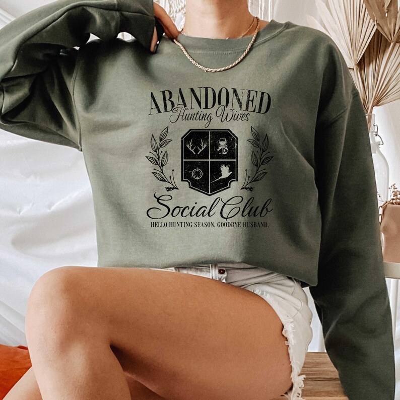 Puede incluir: Una sudadera verde con el texto "Abandoned Hunting Wives Social Club" y un escudo con im&aacute;genes relacionadas con la caza. El texto debajo del escudo dice "Hello Hunting Season, Goodbye Husband."