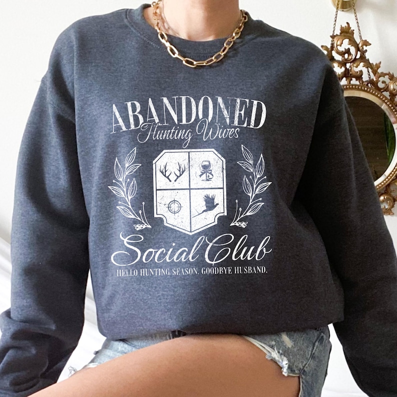 Puede incluir: Una sudadera gris oscura con un estampado gr&aacute;fico blanco que dice "Abandoned Hunting Wives Social Club. Hello Hunting Season, Goodbye Husband."