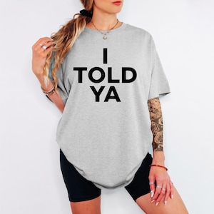 May include: A pale grey t-shirt featuring the phrase "I TOLD YA" in bold black lettering. The top has short sleeves and a loose fit. The t-shirt is paired with black shorts, creating a casual ensemble.