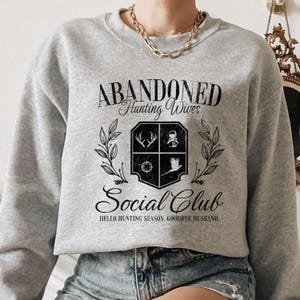 Puede incluir: Sudadera gris con el texto "Abandoned Hunting Wives Social Club" y las palabras "Hello Hunting Season, Goodbye Husband." La sudadera presenta un gr&aacute;fico en blanco y negro de un escudo con s&iacute;mbolos relacionados con la caza.
