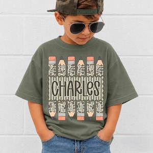Puede incluir: Una camiseta de manga corta color verde oliva con un estampado gráfico de seis lápices verticales con patrón de camuflaje. Los lápices rodean un recuadro central que contiene el nombre CHARLES en una fuente oscura, redondeada y negrita.