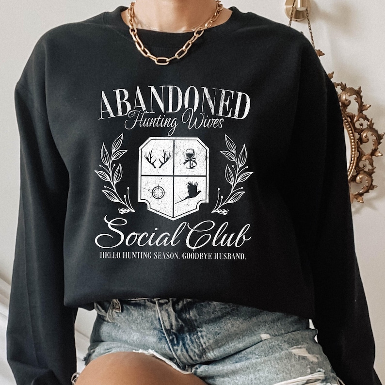 Puede incluir: Sudadera negra con un dise&ntilde;o gr&aacute;fico blanco que dice "Abandoned Hunting Wives Social Club" con un escudo que presenta un ciervo, una copa de vino, un objetivo y un p&aacute;jaro. El texto debajo del escudo dice "Hello Hunting Season, Goodbye Husband."