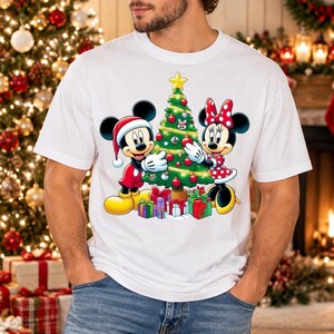 Puede incluir: Una camiseta blanca de manga corta con un gráfico de Mickey Mouse con gorro de Papá Noel y Minnie Mouse con vestido de lunares rojos decorando un árbol de Navidad con adornos y una estrella amarilla, rodeados de cajas de regalo.