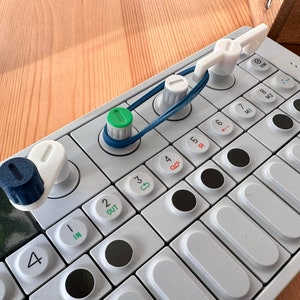 Könnte beinhalten: Eine weiße elektronische Musiktastatur mit schwarzen Tasten und Knöpfen. Die Tastatur hat einen kleinen Bildschirm und eine Reihe weißer Tasten, die mit Zahlen und Buchstaben beschriftet sind. Es gibt auch zwei weiße Knöpfe mit grünen und blauen Akzenten.