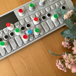 May include: A white and gray electronic music keyboard with red, green, and blue buttons. The keyboard has a small screen and several buttons labeled with numbers and letters. The keyboard is sitting on a wooden surface.