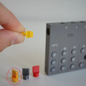 Peut inclure: Gros plan sur quatre boutons en plastique jaune, gris et rouge pour un instrument de musique. Les boutons sont sur une surface blanche. Le texte "OP-Z" est visible sur l'instrument de musique gris en arrière-plan.
