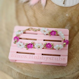 Könnte beinhalten: Ein rosa-weißes Perlenarmband mit goldenen Akzenten. Das Armband wird auf einer rosa Palette mit dem Text "Bines Hobbystube" präsentiert.