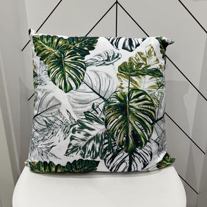 May include: Square decorative pillow with a white background and a repeating pattern of large green and white tropical leaves. The pillow is placed on a white chair. The pillow has a tropical theme.
