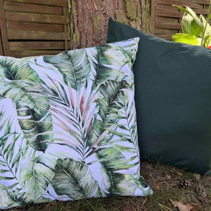 May include: Two decorative throw pillows. One pillow features a vibrant tropical leaf pattern in shades of green, white, and pink. The other pillow is a solid dark green. Both pillows are square and rest on grass near a tree.