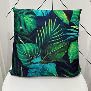 May include: Square decorative pillow with a tropical leaf pattern. The pillow features large green leaves with varying shades of green and teal against a dark blue background. The pillow is placed on a white chair.
