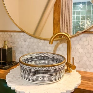 May include: A white ceramic sink with a black and gold geometric pattern, a gold faucet, and a marble countertop.