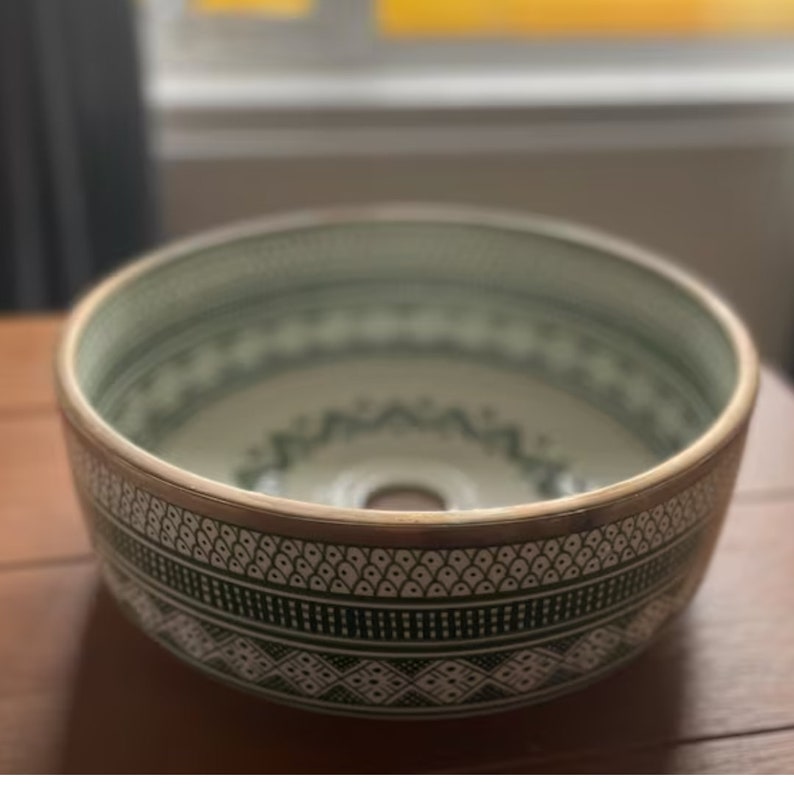 May include: A white ceramic sink with a green and white geometric pattern. The sink has a silver rim and a drain hole in the center.