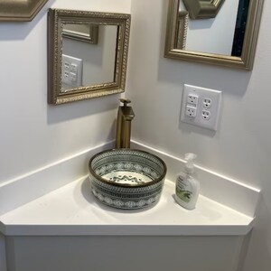 May include: A white ceramic sink with a green and white geometric pattern, a gold faucet, and a bottle of hand soap on a white countertop.