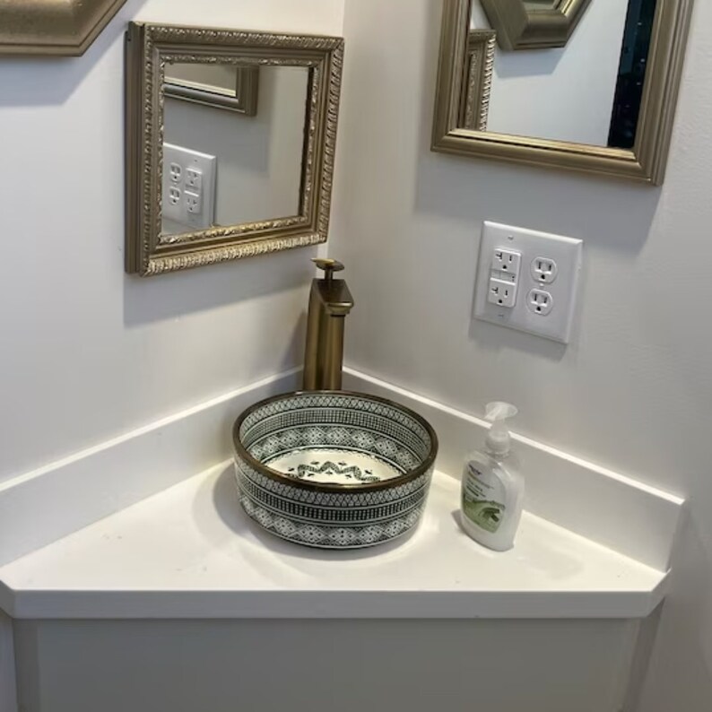 May include: A white ceramic sink with a green and white geometric pattern, a gold faucet, and a bottle of hand soap on a white countertop.