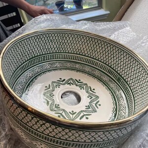 May include: A white ceramic sink with a green and white geometric pattern. The sink has a gold rim and a drain hole in the center.
