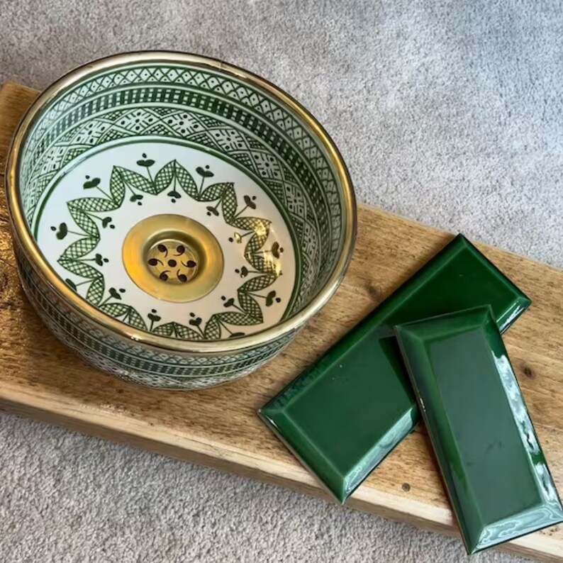May include: A green and white ceramic sink with a gold rim and a brass drain. The sink is on a wooden surface with two green ceramic tiles.