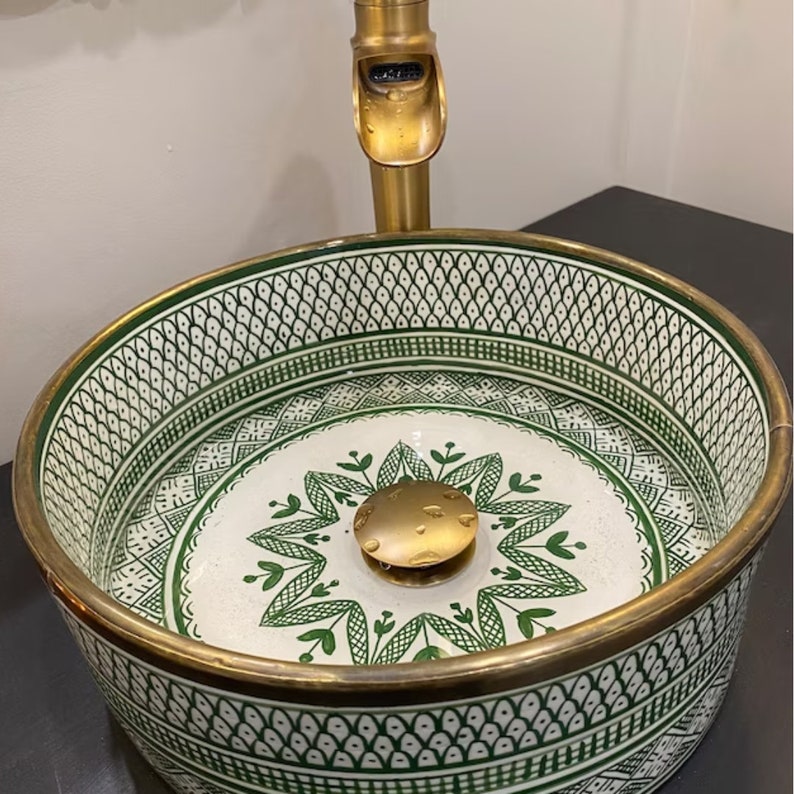 May include: A white ceramic sink with a green and gold geometric pattern. The sink has a brass drain stopper and is designed for a bathroom.
