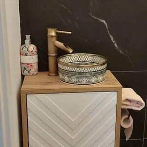 May include: A white ceramic bathroom sink with a black and gold geometric pattern. The sink is on a wooden countertop with a white cabinet with a chevron pattern. A gold faucet and a bottle of hand soap are also visible.