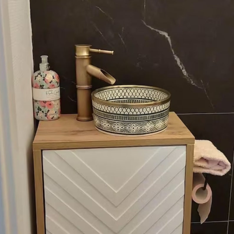 May include: A white ceramic bathroom sink with a black and gold geometric pattern. The sink is on a wooden countertop with a white cabinet with a chevron pattern. A gold faucet and a bottle of hand soap are also visible.
