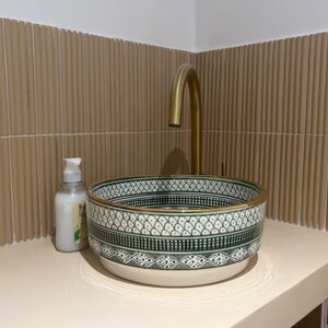May include: A white ceramic sink with a green and black geometric pattern and a gold rim. The sink is mounted on a wall with beige tiles. A gold faucet is mounted above the sink.