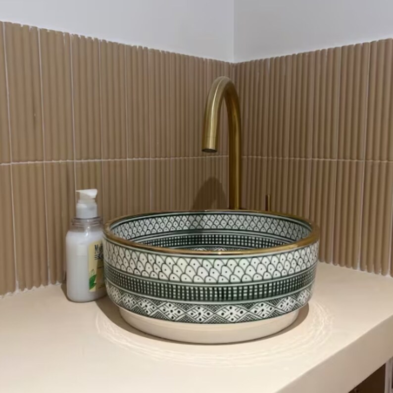 May include: A white ceramic sink with a green and black geometric pattern and a gold rim. The sink is mounted on a wall with beige tiles. A gold faucet is mounted above the sink.