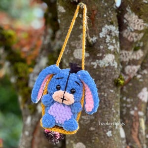 May include: A handmade blue plush toy of a donkey character with pink inner ears, a black hat, and a light purple belly. The toy is suspended by a yellow strap, and the background is a blurred tree.