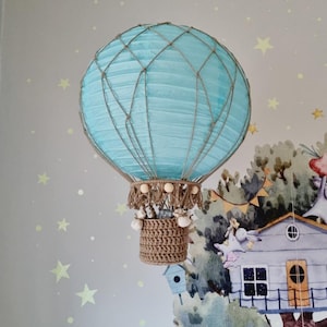 Puede incluir: Un globo aerostático azul claro hecho de papel y cuerda con una cesta de crochet marrón colgando. El globo está decorado con pequeñas cuentas blancas y está colgado del techo.