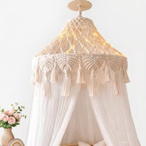May include: A cream macrame canopy bed with a sheer white curtain. The canopy features tassels and fairy lights. The bed has a rattan frame and is decorated with pillows and a cloud-shaped cushion.