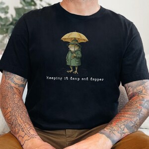 May include: Black t-shirt featuring a frog character wearing a green raincoat and holding a yellow umbrella. The text "Keeping it damp and dapper" is printed below the frog. The shirt is worn by a person with tattoos.