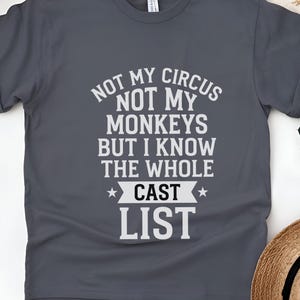 May include: A dark gray short-sleeved t-shirt featuring white text that reads: NOT MY CIRCUS NOT MY MONKEYS BUT I KNOW THE WHOLE CAST LIST. The shirt has a crew neckline and a standard fit, with the graphic centered on the front of the garment.