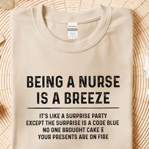 May include: Sand-colored t-shirt with the text "BEING A NURSE IS A BREEZE." The shirt also has the text "IT'S LIKE A SURPRISE PARTY EXCEPT THE SURPRISE IS A CODE BLUE NO ONE BROUGHT CAKE & YOUR PRESENTS ARE ON FIRE."