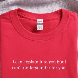 May include: A bright red crew neck t-shirt featuring the white printed text "i can explain it to you but i can't understand it for you." The shirt has a white brand label at the inner neckline and is folded neatly on a soft, textured fabric surface.