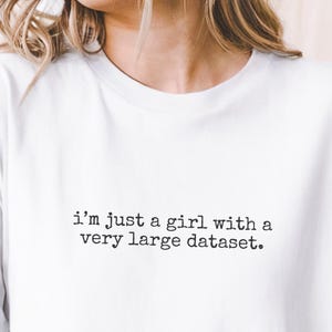 May include: White t-shirt featuring the phrase "I'm just a girl with a very large dataset." The shirt has a casual style with short sleeves. The text is printed in a plain, sans-serif font.