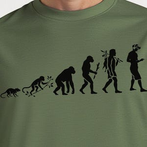 Może przedstawiać: Zielona koszulka w kolorze wojskowym z czarną grafiką przedstawiającą ewolucję człowieka, od małpy do współczesnej osoby z zestawem VR. Koszulka jest w jednolitym kolorze z okrągłym dekoltem. Tekst "Military Green" znajduje się w okręgu.