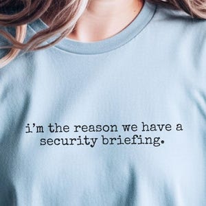 Puede incluir: Camiseta azul claro con la frase "i'm the reason we have a security briefing." El texto está en una fuente sencilla. La camiseta es de color azul claro liso.
