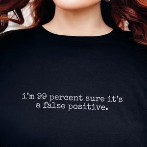 May include: Black t-shirt with the text "i'm 99 percent sure it's a false positive" in white. The shirt is a crew neck style. The person in the image is holding their hair.