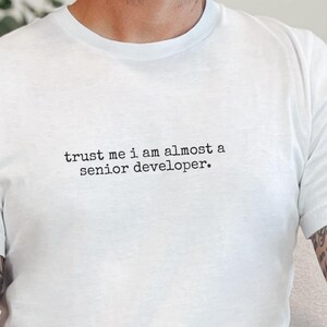 May include: A white short-sleeved t-shirt featuring the black typewriter-style text "trust me i am almost a senior developer." printed across the chest. The garment has a crew neckline and a standard fit, providing a clean and simple appearance.