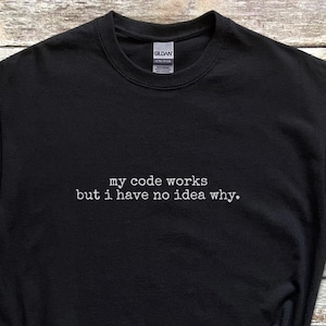 May include: Black t-shirt with the text "my code works but I have no idea why." The shirt is paired with blue denim shorts, black trainers, a straw hat, a smartwatch, and wireless earbuds. The items are arranged on a wooden surface.