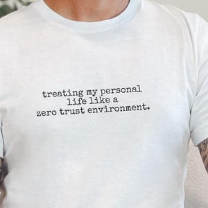 May include: A white short-sleeved t-shirt featuring black typewriter-style text printed on the chest that reads, "treating my personal life like a zero trust environment." The shirt has a crew neckline and a standard fit.