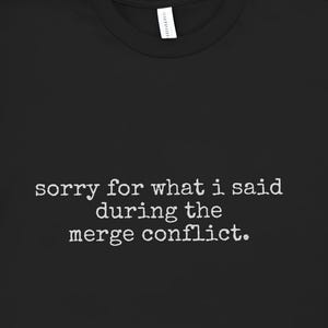 May include: Black t-shirt with the text "sorry for what i said during the merge conflict" in a white, simple font. The shirt is displayed on a white surface with a succulent plant and a straw hat in the background.