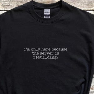 Puede incluir: Camiseta negra con el texto "I'm only here because the server is rebuilding." La camiseta se combina con pantalones cortos de mezclilla azules, zapatillas negras, un sombrero de paja, un reloj inteligente y auriculares inalámbricos. Los artículos están colocados sobre una superficie de madera.