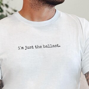 May include: White crew neck t-shirt with the text "I'm just the ballast." The shirt is made of a soft material. The person is wearing brown pants. The person has tattoos on their arms.