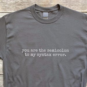 May include: A charcoal gray t-shirt with the text "you are the semicolon to my syntax error." The shirt is paired with blue denim shorts, black sneakers, a straw hat, and a smartwatch.