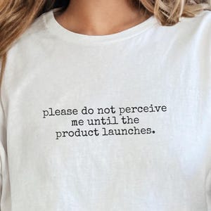 May include: A white t-shirt with the text "please do not perceive me until the product launches" in black. The shirt is worn with blue jeans. The overall theme is casual wear.