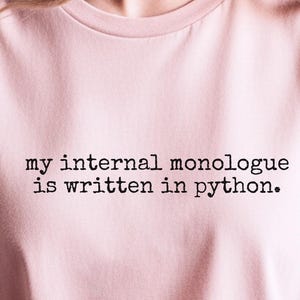 May include: A light pink short-sleeved t-shirt featuring the black printed text 'my internal monologue is written in python.' The shirt has a crew neckline and a standard fit, with a small circular label in the corner indicating the color.