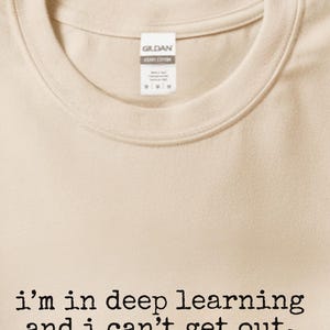 May include: A sand-colored crew neck t-shirt featuring the black text 'i'm in deep learning and i can't get out.' printed in a typewriter-style font on the chest. The shirt has a Gildan brand label visible at the inner neckline.