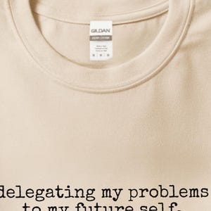 May include: A sand-colored crew neck t-shirt featuring the black printed text 'delegating my problems to my future self.' The shirt has a Gildan brand label at the inner neckline and is displayed in a flat, folded orientation.