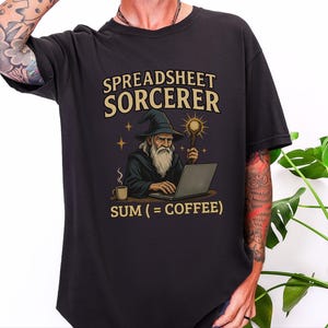 May include: A black t-shirt featuring a graphic of a wizard with a long white beard and hat working on a laptop. The text reads SPREADSHEET SORCERER above the wizard and SUM (= COFFEE) below, with a coffee mug and a glowing staff visible.