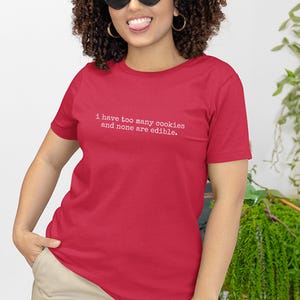 Könnte beinhalten: Ein rotes T-Shirt mit dem weißen Text "I have too many cookies and none are edible." Das Shirt ist einfarbig und hat kurze Ärmel. Die Person trägt eine Sonnenbrille und goldene Creolen.